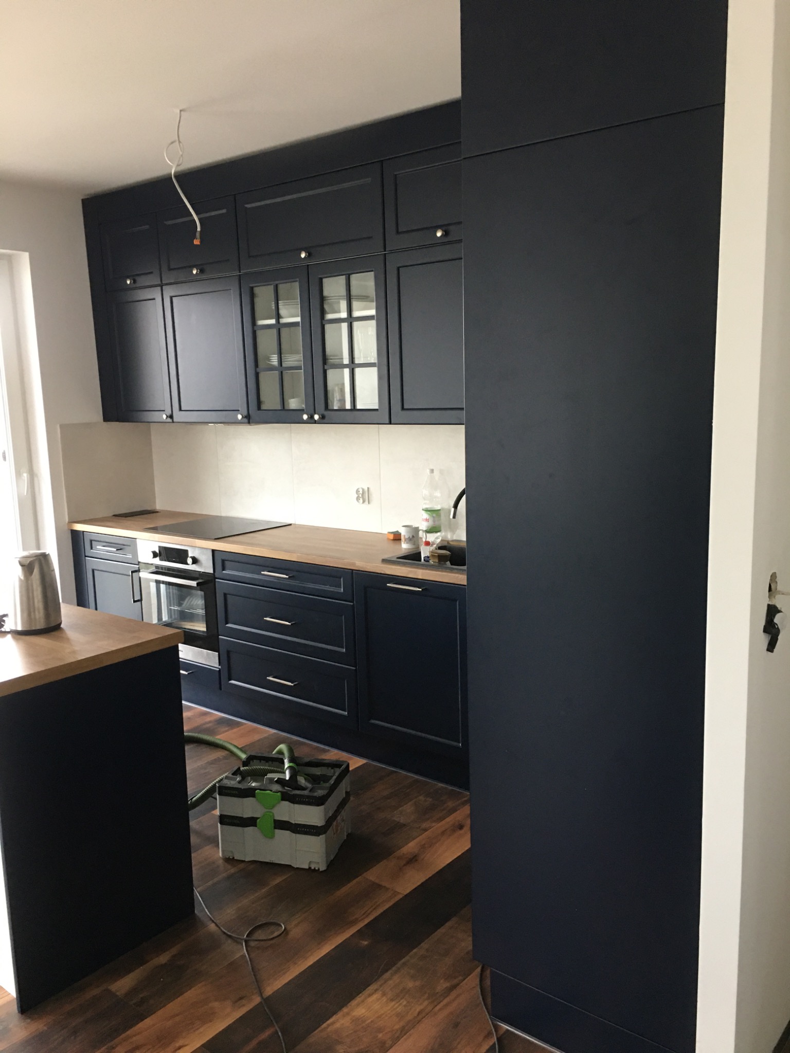 Nowoczesna, granatowa kuchnia z drewnianymi blatami, widoczny odkurzacz budowlany na podłodze podczas montażu.