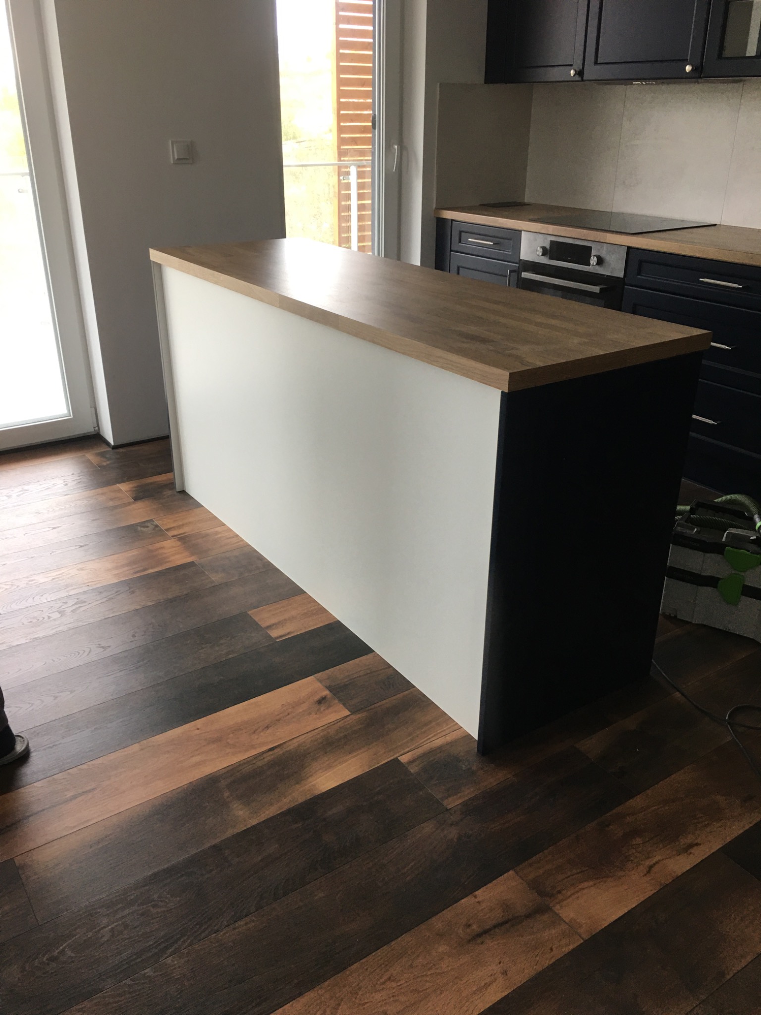 Wyspa kuchenna z blatem imitującym drewno, białą zabudową od frontu i ciemnogranatowymi bokami, na tle ciemnej drewnianej podłogi i zabudowy kuchennej w tym samym kolorze.