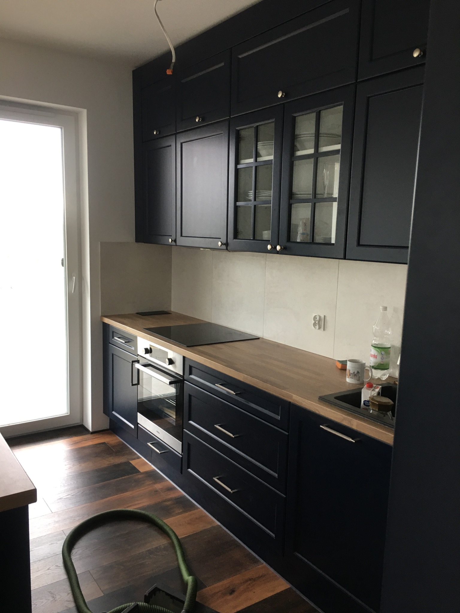 Ciemna zabudowa kuchenna z drewnianym blatem, widoczne szklane witryny górnych szafek, na podłodze panele imitujące drewno, w tle białe ściany i okno, na blacie kuchennym zlewozmywak, płyta...