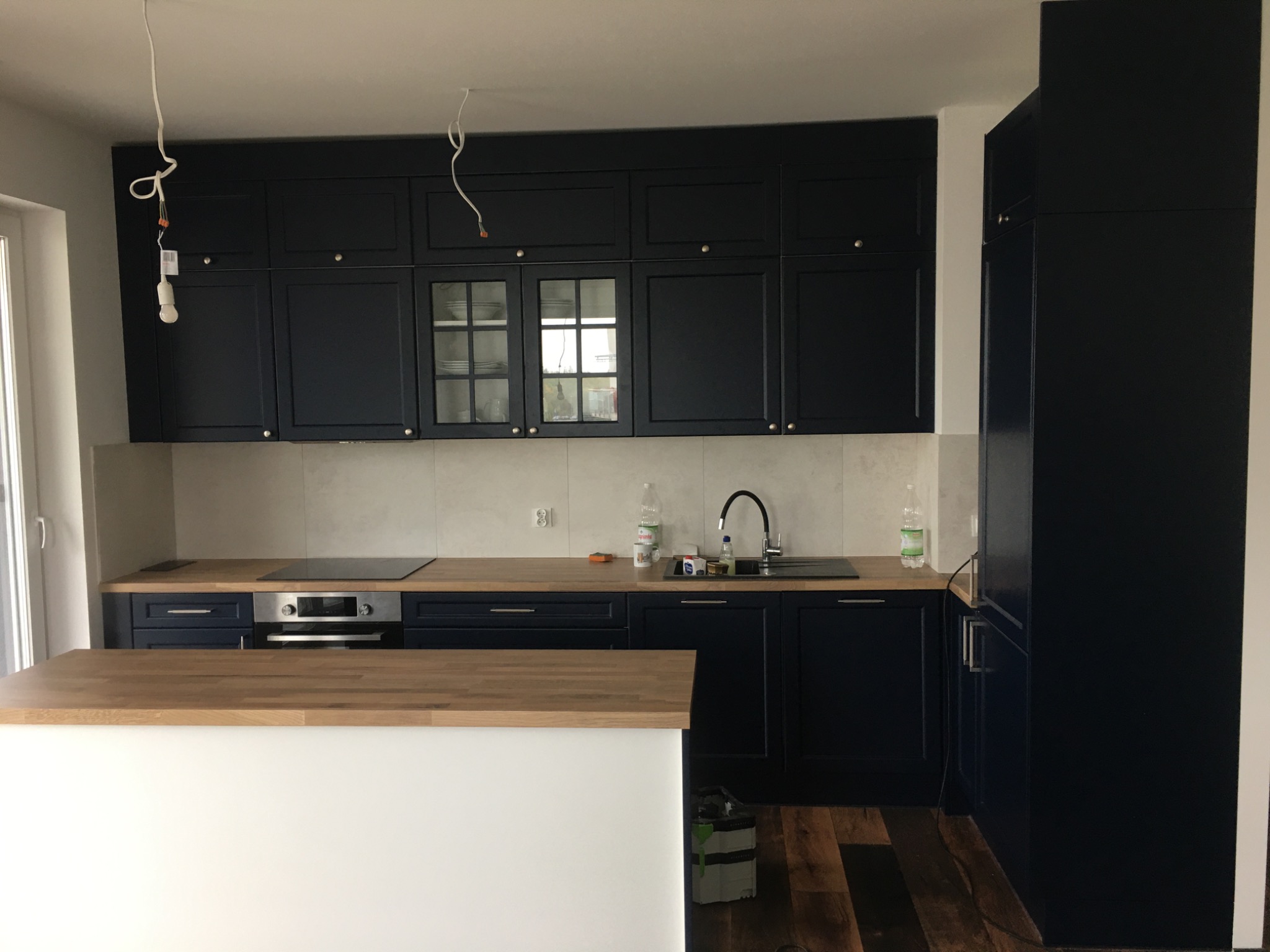 Nowoczesna, granatowa kuchnia z drewnianym blatem, wyspą kuchenną z białym frontem i niedokończonym oświetleniem sufitowym.