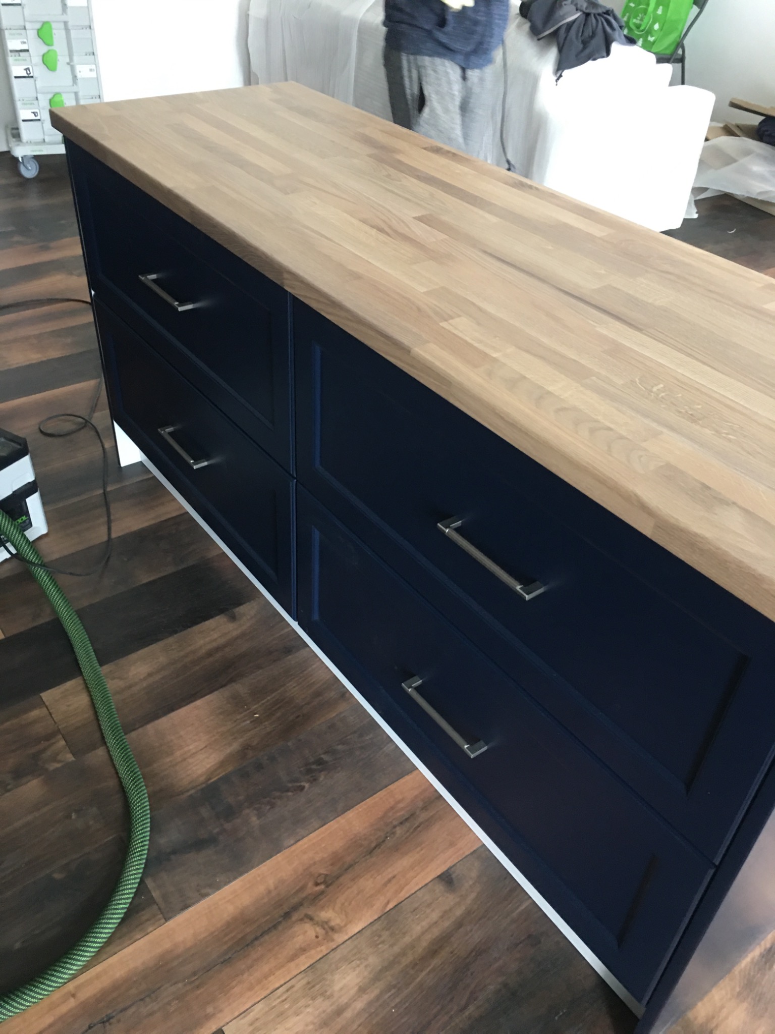 Niebieska szafka kuchenna z drewnianym blatem, widoczne metalowe uchwyty szuflad, na podłodze leży wąż od odkurzacza, w tle osoba i meble przykryte folią.