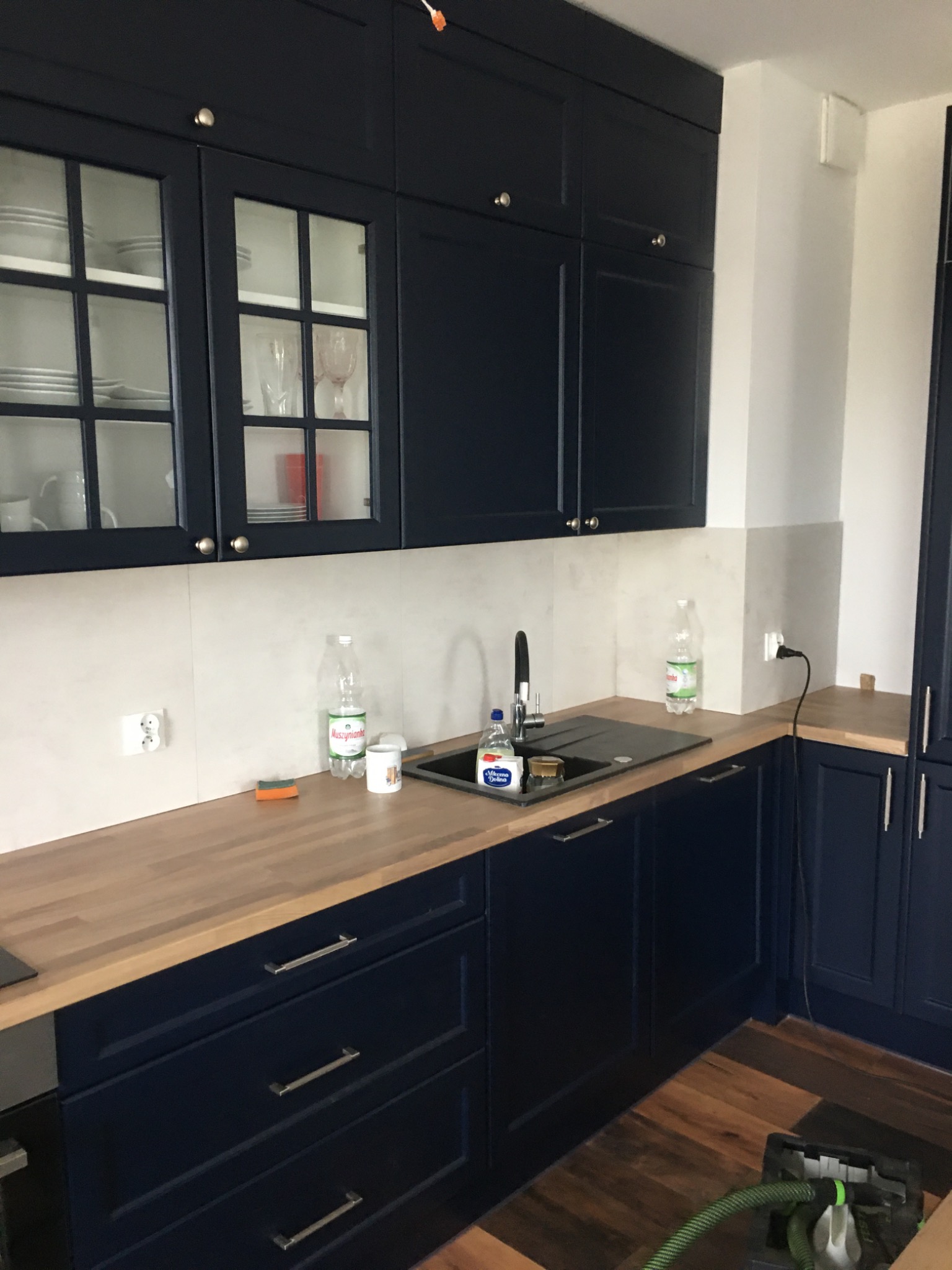 Nowoczesna, granatowa zabudowa kuchenna z drewnianym blatem, szklane witryny w szafkach wiszących, widoczne naczynia, czarny zlewozmywak, na blacie butelki wody mineralnej i płyn do naczyń, podłoga...