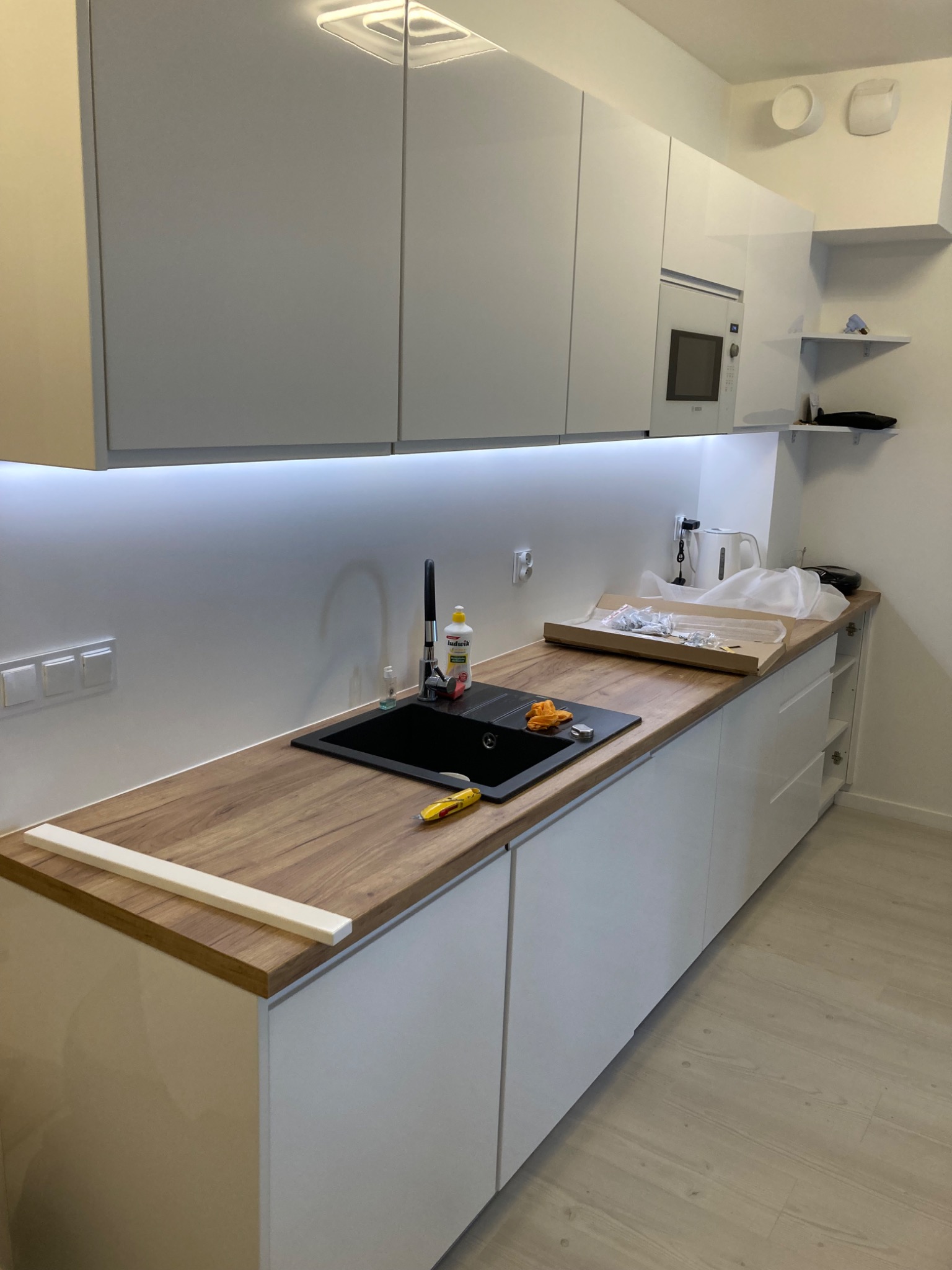 Nowoczesna, biała kuchnia z drewnianym blatem, czarnym zlewozmywakiem i podświetleniem LED pod szafkami, w trakcie montażu, widoczny fragment listwy i narzędzie na blacie.