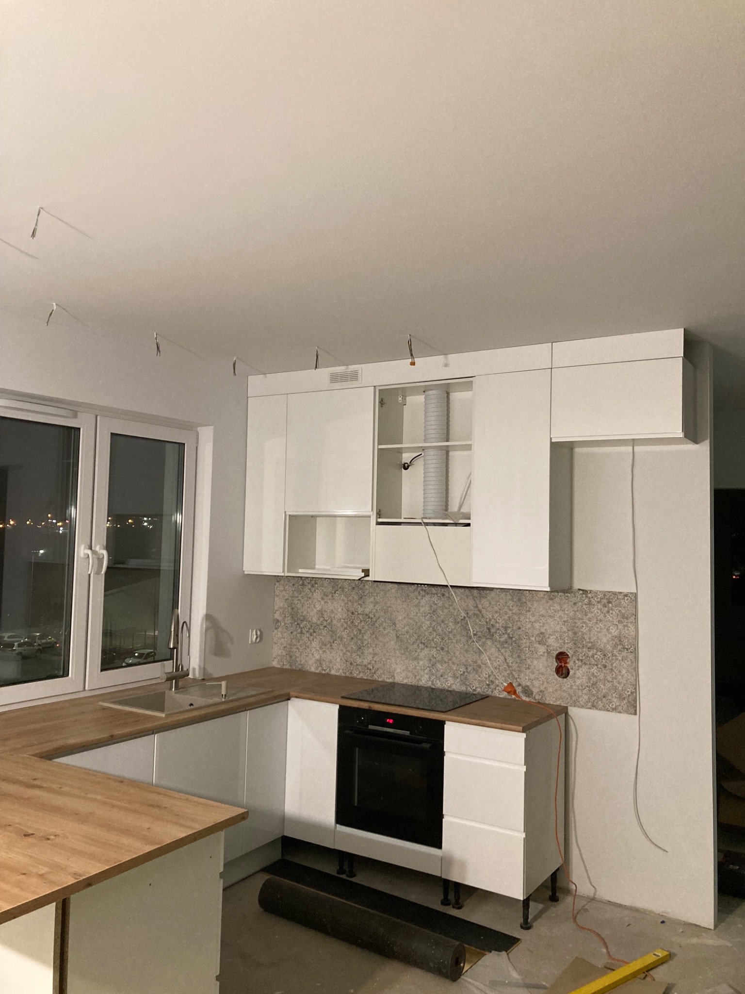 Wnętrze w trakcie montażu: białe szafki kuchenne z drewnianym blatem, widoczne przewody elektryczne, niedokończone płytki na ścianie i rozłożony zwój podłogi w tle okna.