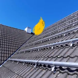 Montaż szyn pod panele fotowoltaiczne na dachu z ciemnej dachówki karpiówki, widoczne mocowania i fragment sąsiedniego dachu z kominem wentylacyjnym na tle błękitnego nieba.