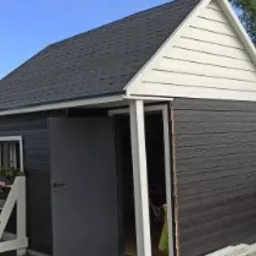 Niewielka, ciemna budowla z dwuspadowym dachem pokrytym ciemną dachówką bitumiczną, z otwartymi drzwiami i białym wykończeniem szczytu, na tle zieleni ogrodu i domu z czerwoną dachówką.