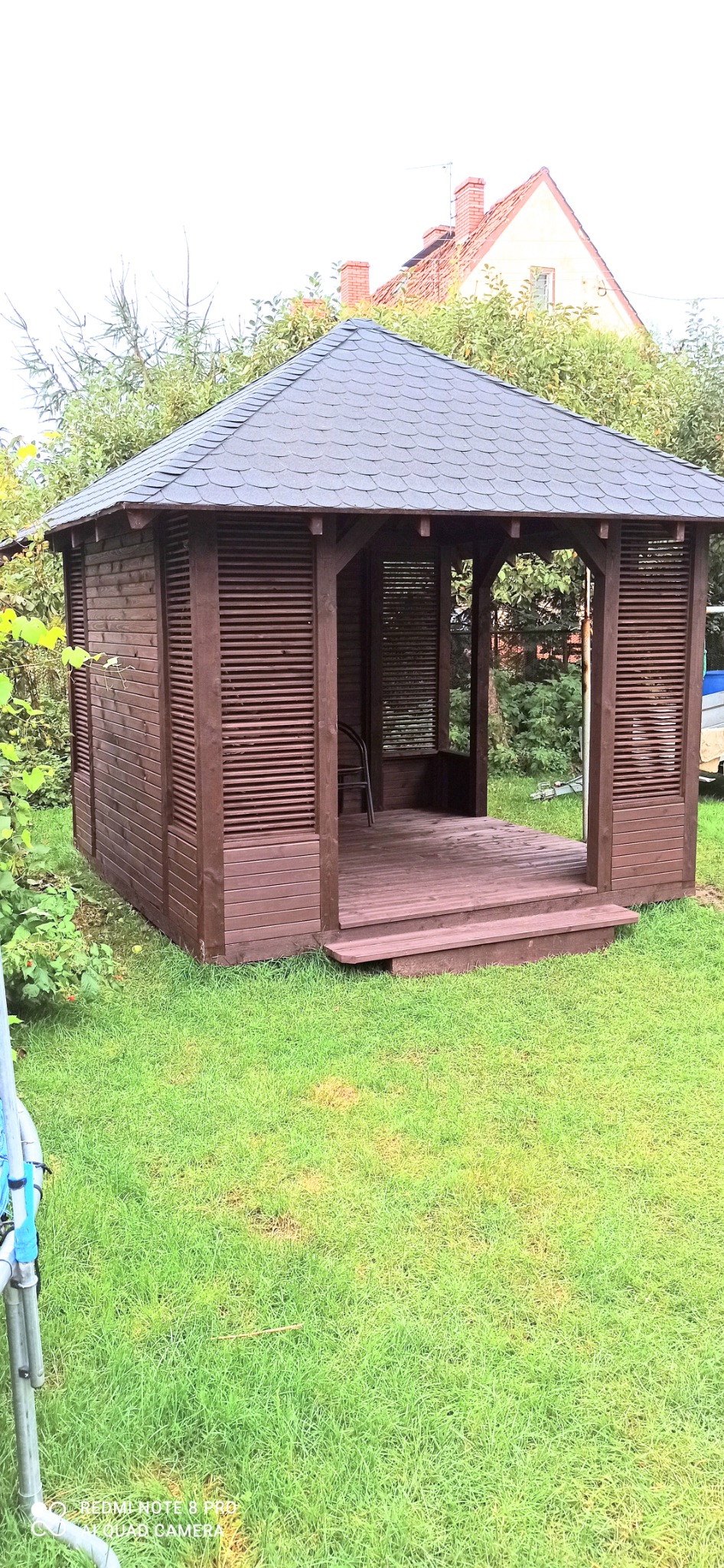 Brązowa altana ogrodowa z drewnianą podłogą i szarym dachem z gontów bitumicznych, otoczona zielonym trawnikiem i drzewami.