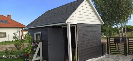 Niewielka, ciemna budowla z dwuspadowym dachem pokrytym ciemną dachówką bitumiczną, z otwartymi drzwiami i białym wykończeniem szczytu, na tle zieleni ogrodu i domu z czerwoną dachówką.