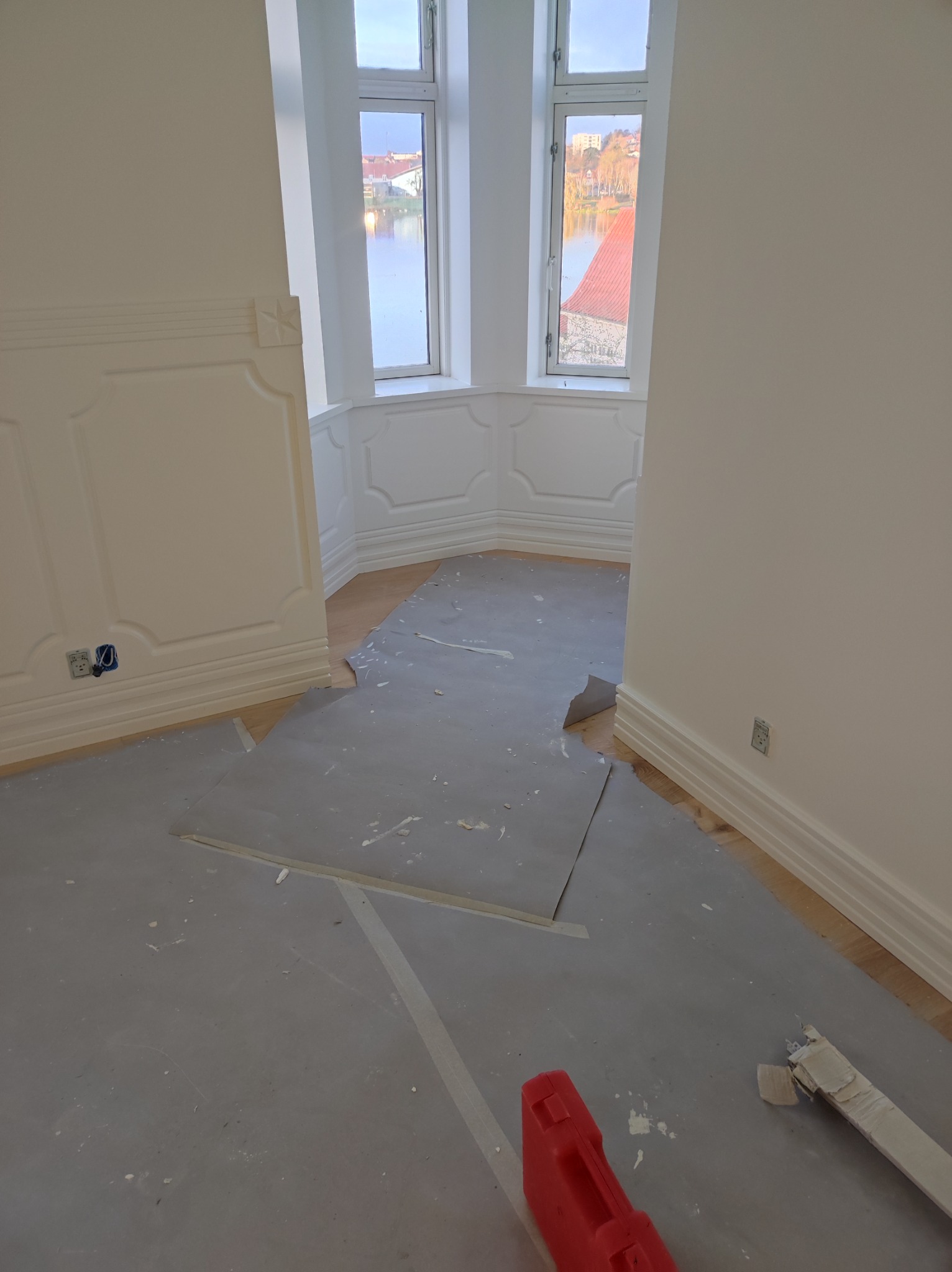 Przygotowanie do ułożenia podłogi: zabezpieczona powierzchnia, widoczne listwy przypodłogowe i skrzynka narzędziowa, w tle okno z widokiem na jezioro.