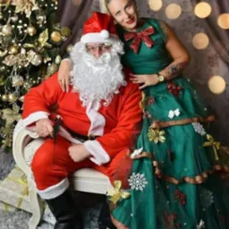 Animator w przebraniu Świętego Mikołaja siedzi na krześle obok kobiety w zielonej sukience przypominającej choinkę, na tle świątecznej dekoracji z jasnymi punktami świetlnymi.