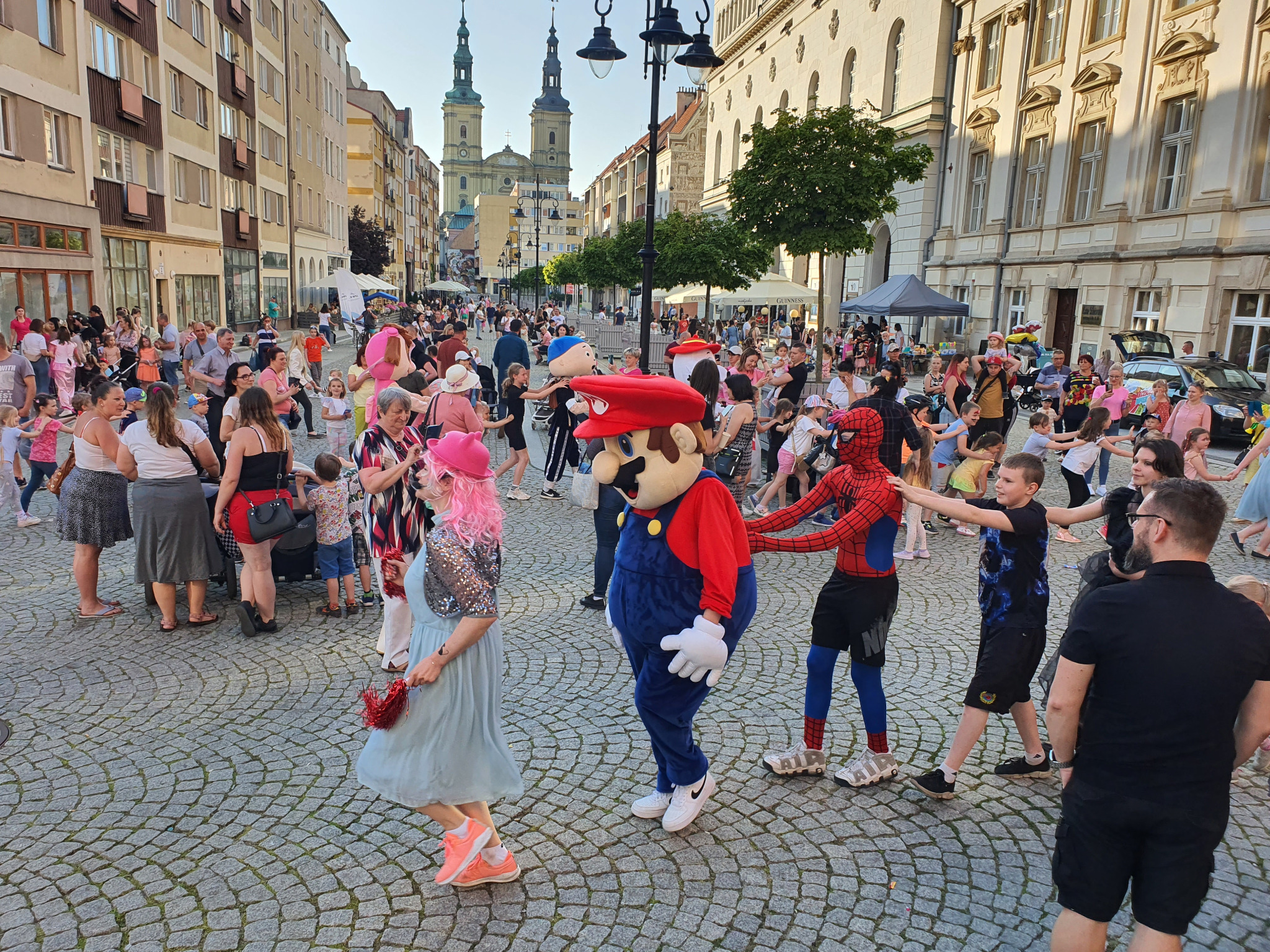 Ulica w mieście z kostki brukowej, tłum ludzi, w tym osoby przebrane za Mario i Spidermana, oraz kobieta w różowej peruce i jasnoniebieskiej sukience, w tle budynki i wieże kościoła.