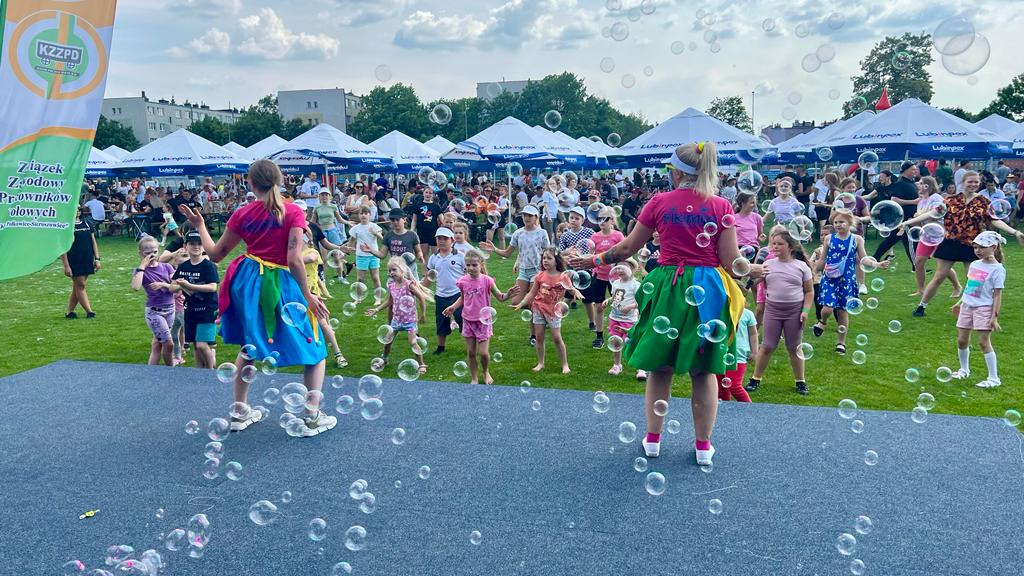 Dwie kobiety w kolorowych strojach stoją na scenie, tyłem do widza, otoczone bańkami mydlanymi, przed dużą grupą dzieci i dorosłych na trawniku, w tle białe namioty i budynki.