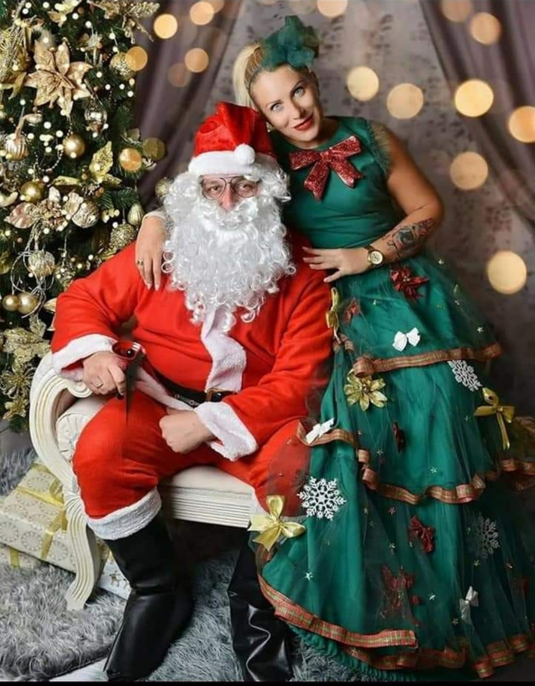 Animator w przebraniu Świętego Mikołaja siedzi na krześle obok kobiety w zielonej sukience przypominającej choinkę, na tle świątecznej dekoracji z jasnymi punktami świetlnymi.