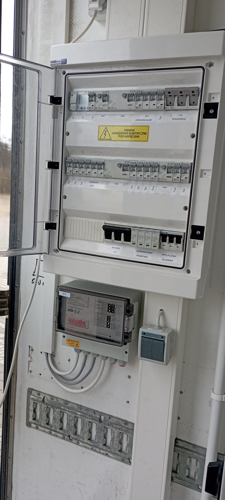Otwarta skrzynka elektryczna z bezpiecznikami, wyłącznikami i modułem alarmowym gazex MD-2.Z, widoczne okablowanie i tabliczka ostrzegawcza o napięciu.