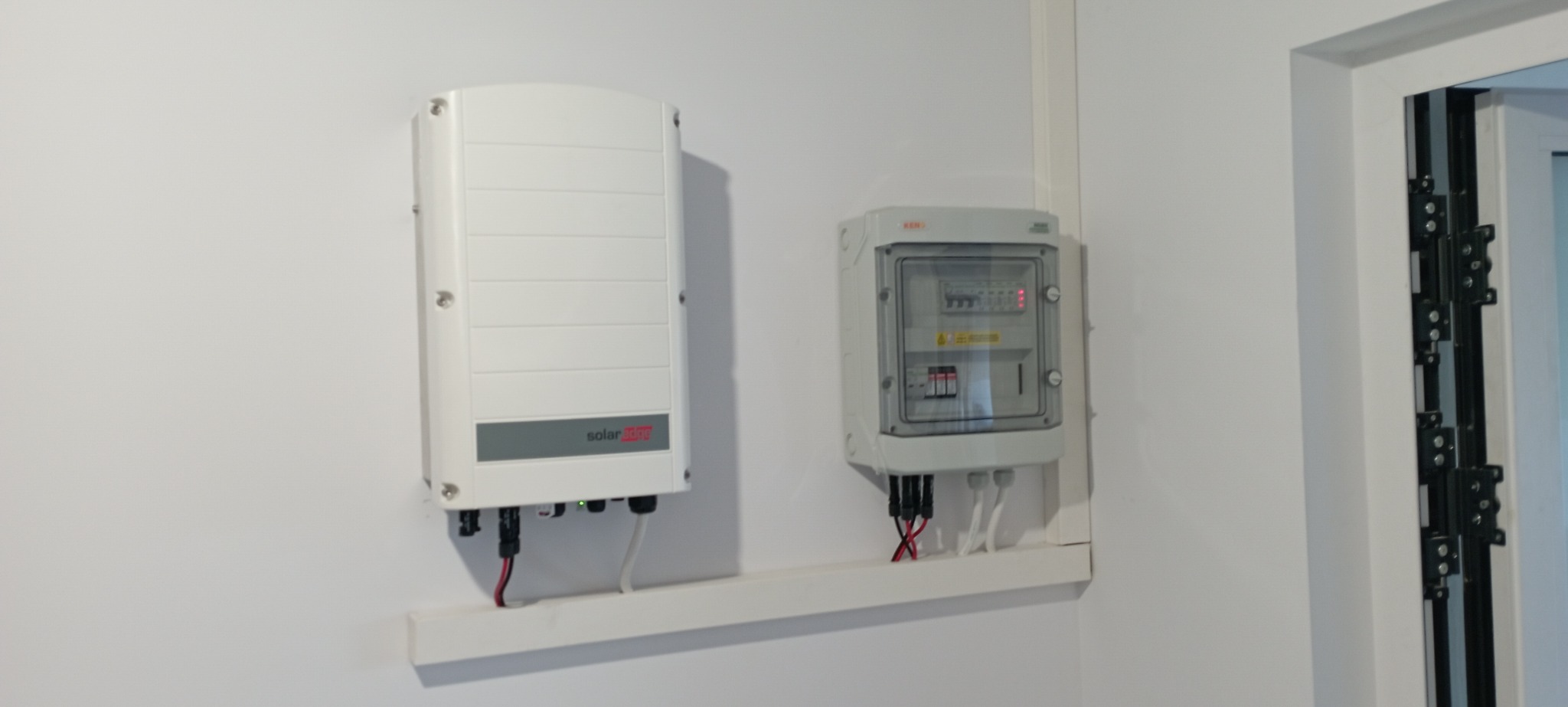 Biała ściana z zamontowanym falownikiem solarnym z logo SolarEdge oraz skrzynką bezpiecznikową z przezroczystą obudową, połączone listwą maskującą.