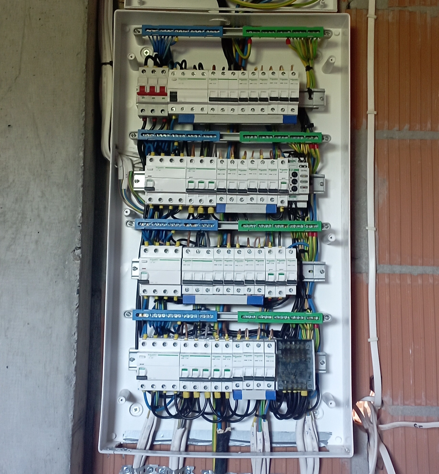 Rozdzielnica elektryczna z zamontowanymi zabezpieczeniami, widoczne kable w kolorach niebieskim, czarnym, zielono-żółtym, umieszczona na ścianie z cegły dziurawki.