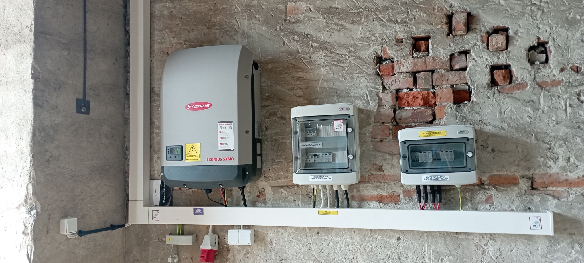 Szary inwerter Fronius Symo zamontowany na ścianie z widoczną cegłą, obok dwie skrzynki rozdzielcze PV DC i AC, kable w białej listwie przypodłogowej.