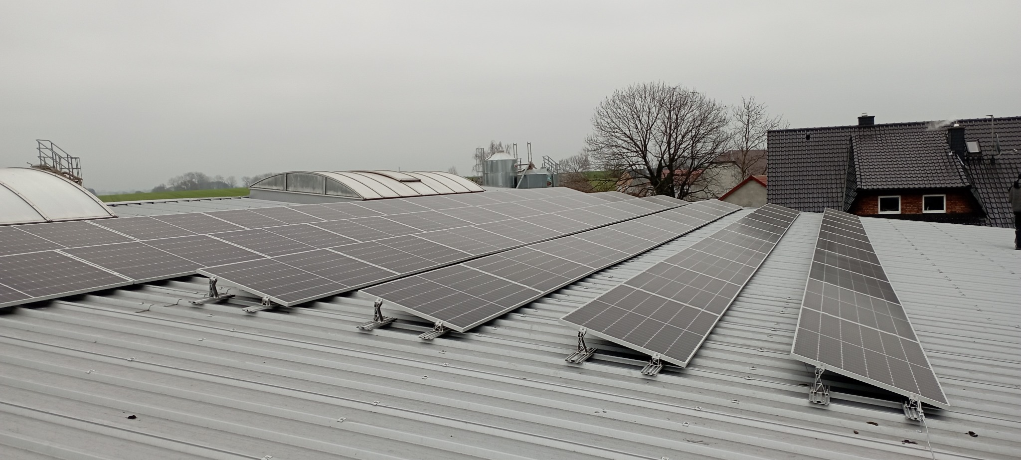 Rząd paneli słonecznych zamontowanych na metalowym dachu przemysłowego budynku w pochmurny dzień, z widocznymi elementami mocującymi i budynkami w tle.