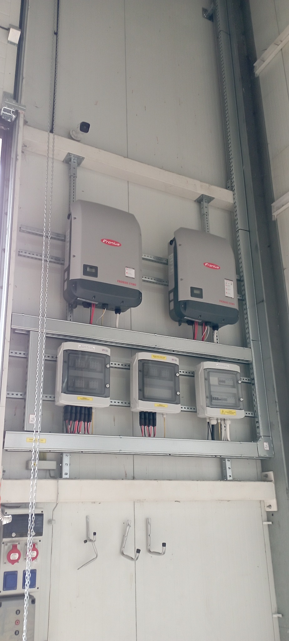 Dwa inwertery Fronius Symo i trzy skrzynki rozdzielcze zamontowane na metalowych szynach w pomieszczeniu przemysłowym, widoczne przewody i elementy instalacji.