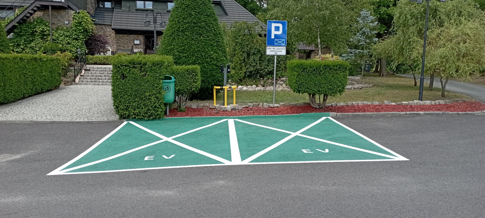 Oznakowane, zielone miejsce parkingowe dla pojazdów elektrycznych (EV) z białymi liniami i napisem, widoczne słupki do ładowania, kosz na śmieci i znak informacyjny na tle zadbanego ogrodu i budynku.