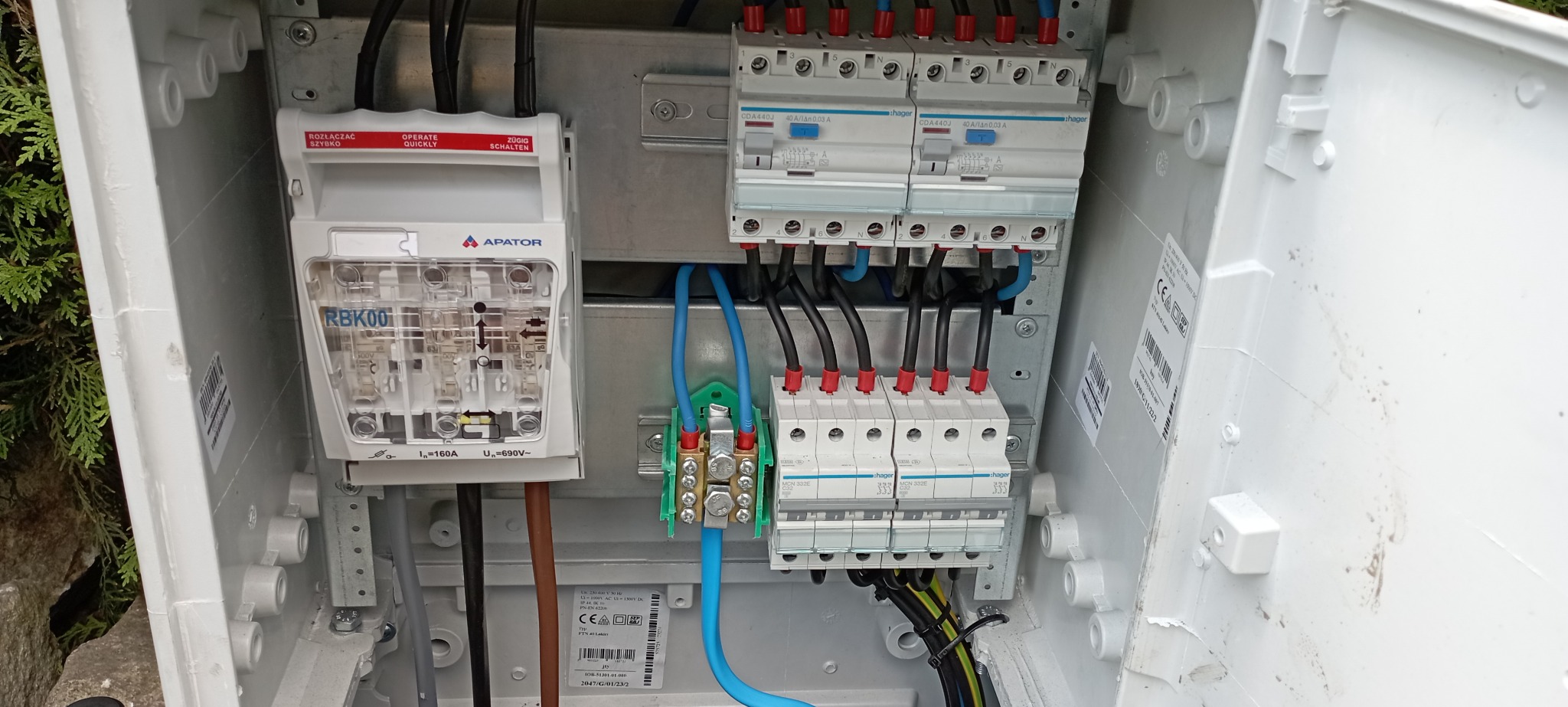 Wnętrze skrzynki elektrycznej z zamontowanymi bezpiecznikami, rozłącznikiem bezpiecznikowym RBK00 firmy Apator, oraz przewodami w kolorach czarnym, niebieskim i żółto-zielonym, widoczne oznaczenia...