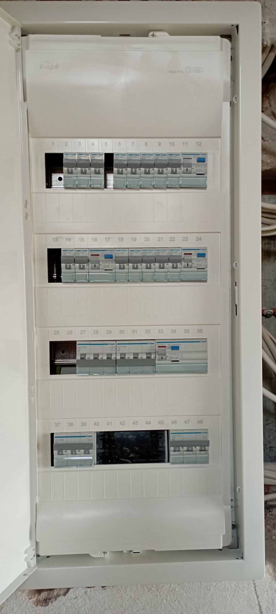 Otwarta, biała skrzynka elektryczna firmy Hager z zainstalowanymi bezpiecznikami i numeracją od 1 do 48, widoczna na tle ściany w trakcie remontu.