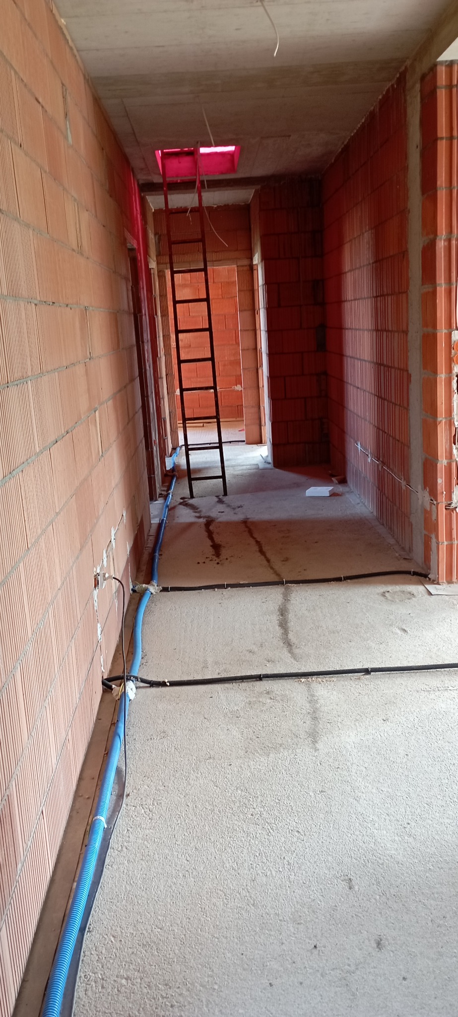 Surowy stan budynku w trakcie prac instalacyjnych: widoczne przewody elektryczne w korytarzu z ceglanymi ścianami i drabiną prowadzącą do otworu w suficie.