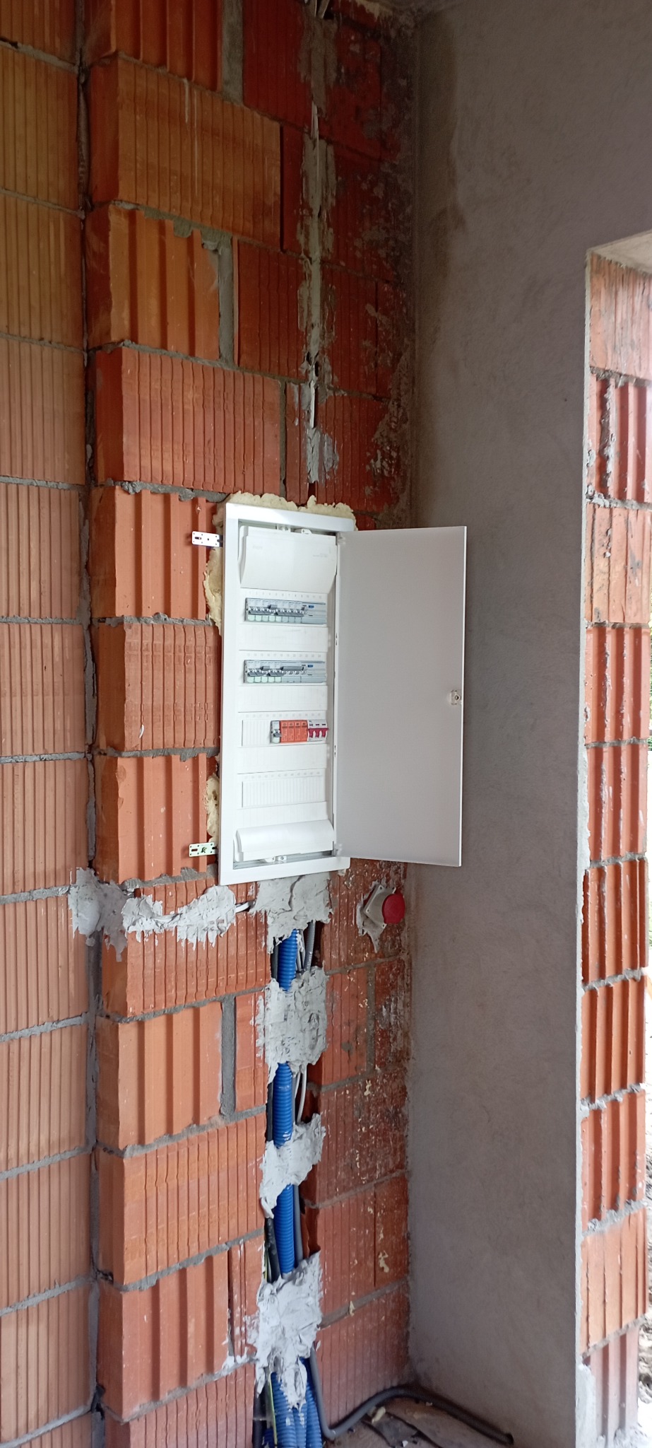 Otwarta skrzynka bezpiecznikowa zamontowana w surowej ścianie z cegieł, widoczne niebieskie rurki instalacyjne i okablowanie w trakcie montażu.