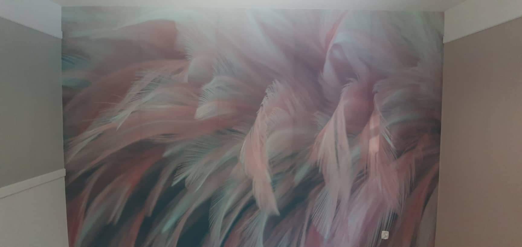 Fragment ściany z fototapetą przedstawiającą abstrakcyjny wzór piór w odcieniach różu i bieli, widoczny fragment listwy przypodłogowej i sąsiedniej ściany pomalowanej na beżowo.