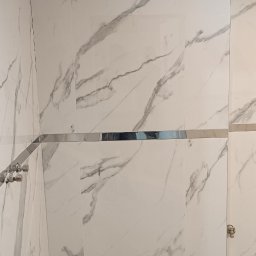 Franciszek Balak - Ściany łazienki wyłożone białymi płytkami imitującymi marmur z szarymi żyłami, widoczne chromowane listwy, otwór w ścianie i niedokończony sufit.