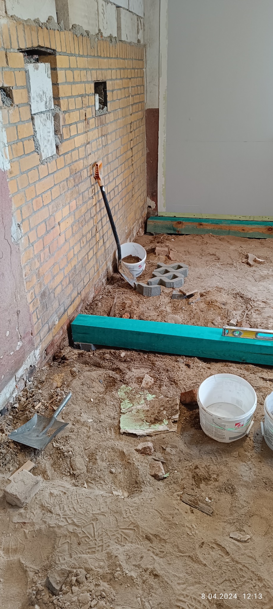 Pokój w trakcie remontu: ściana z cegły z widocznymi ubytkami, podłoga pokryta piaskiem i gruzem, widoczne narzędzia (szpachla, poziomica, łopata) i wiaderka z zaprawą. Fragment ściany pomalowany...