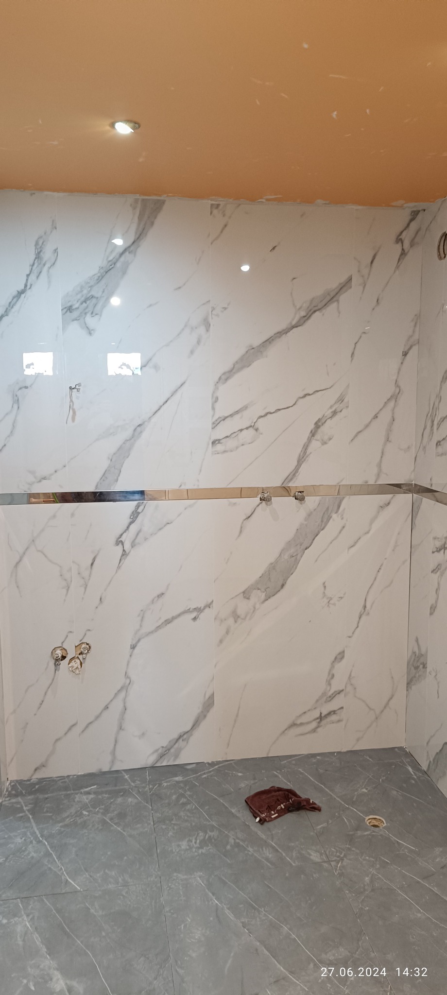 Wnętrze łazienki w trakcie prac wykończeniowych: marmurowe płytki na ścianach z widocznymi otworami na armaturę, szare płytki na podłodze, pomarańczowy sufit z zamontowanym oświetleniem punktowym...