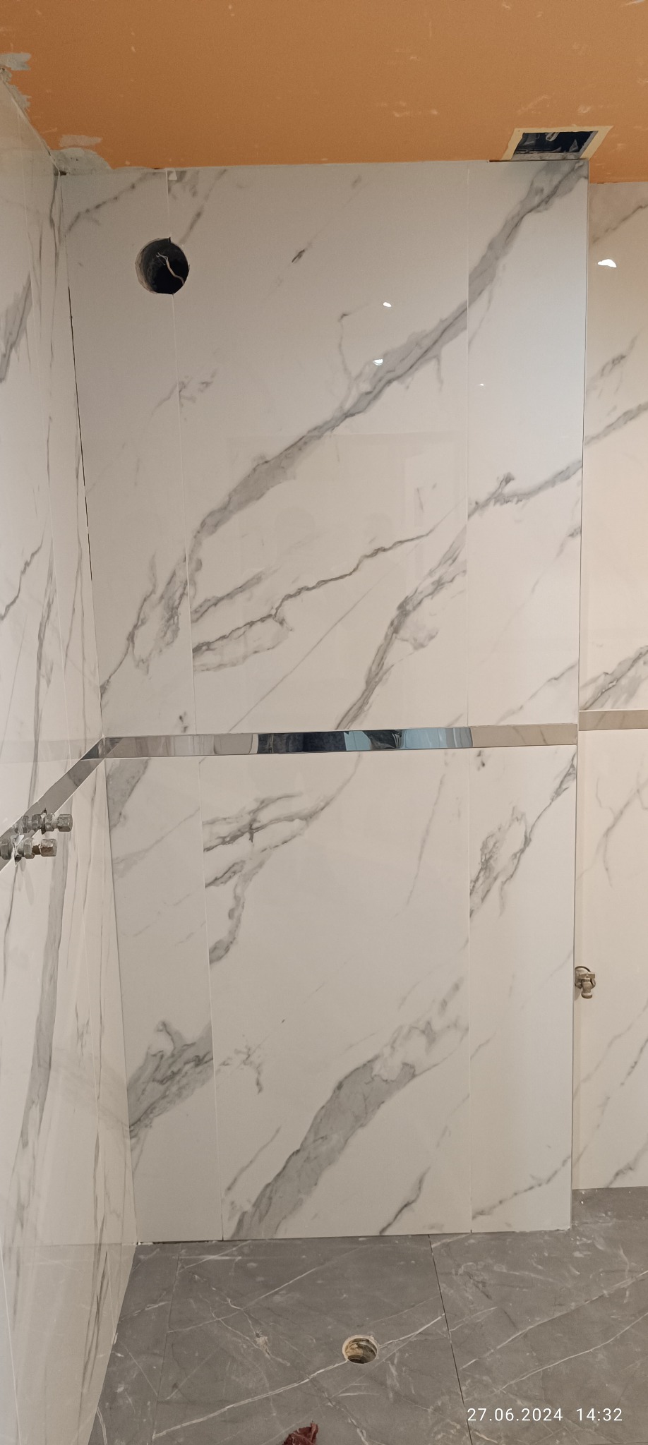 Ściany łazienki wyłożone białymi płytkami imitującymi marmur z szarymi żyłami, widoczne chromowane listwy, otwór w ścianie i niedokończony sufit.