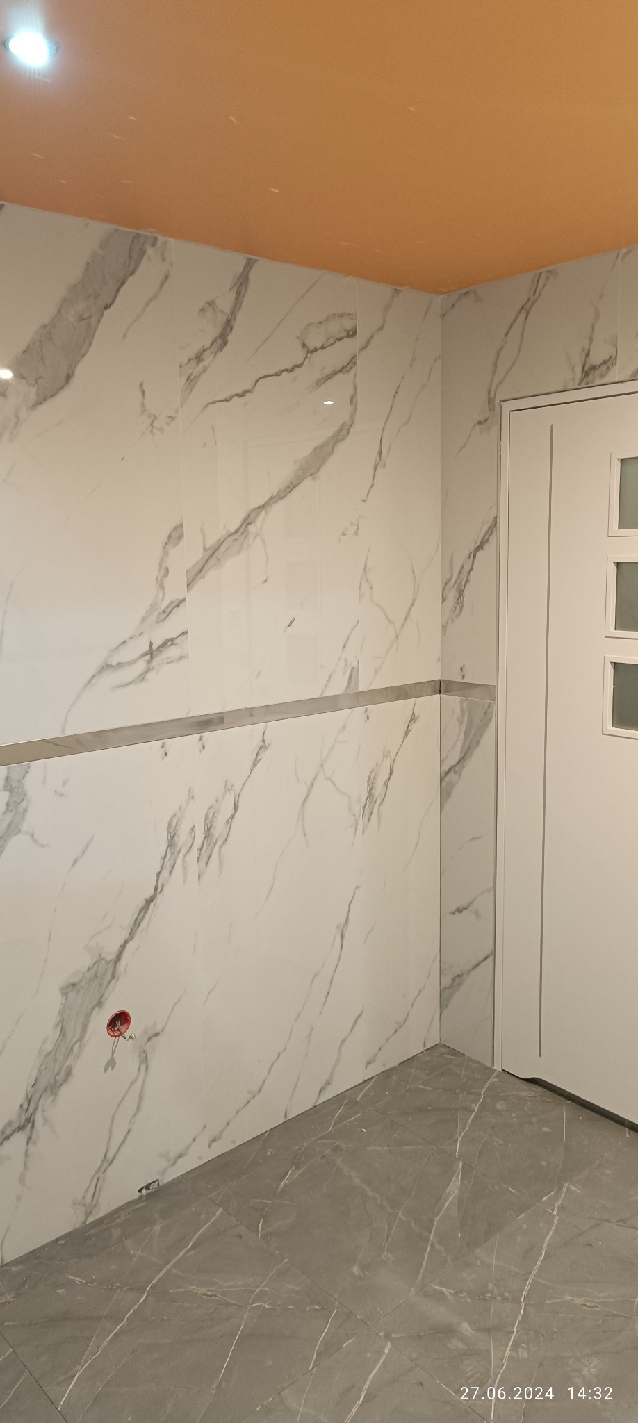 Wykończona łazienka z białymi płytkami imitującymi marmur na ścianach i szarymi płytkami na podłodze, widoczne gniazdko elektryczne i pomarańczowy sufit.