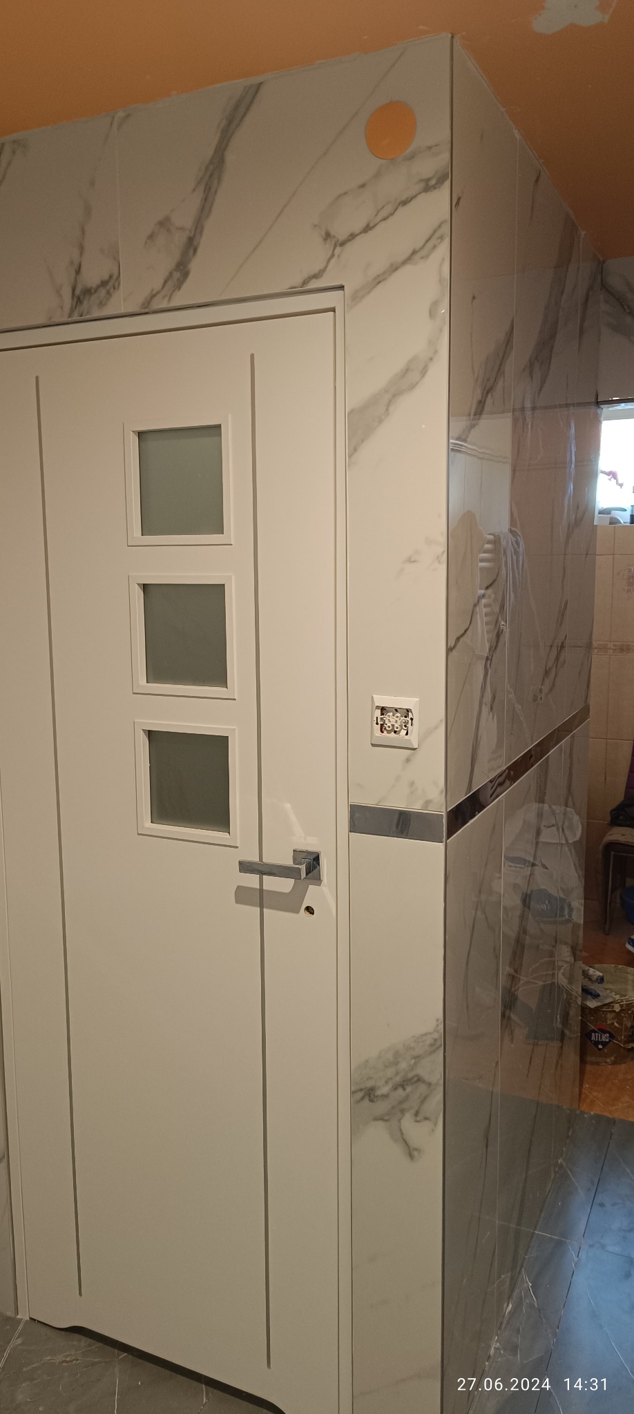 Wykończona łazienka z białymi płytkami imitującymi marmur, białe drzwi z trzema kwadratowymi okienkami, wystający przewód elektryczny ze ściany, pomarańczowy sufit i widoczne wiadro z zaprawą.