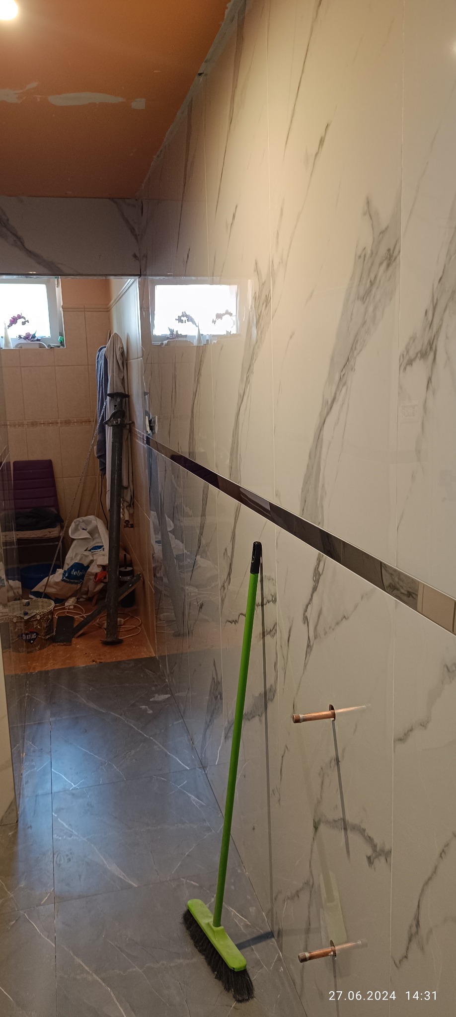 Fragment łazienki w trakcie remontu: marmurowe płytki na ścianach, szare płytki na podłodze, widoczne rury instalacji wodnej oraz stojąca zielona zmiotka, w tle widoczne narzędzia i materiały...