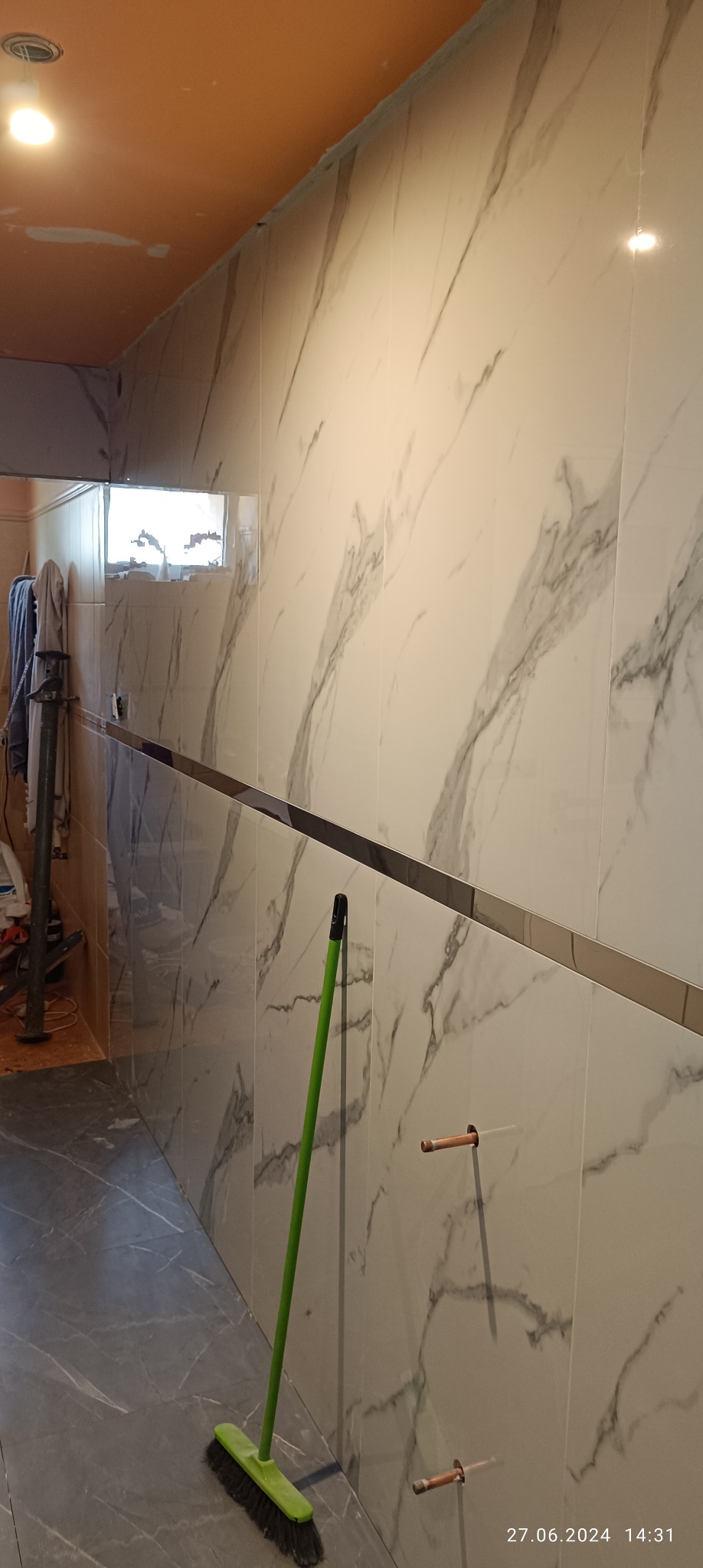 Wnętrze łazienki w trakcie prac wykończeniowych: widoczne marmurowe płytki na ścianach z metalową listwą dekoracyjną, przygotowane otwory na instalacje wodne, szara podłoga z wzorem oraz zielona...