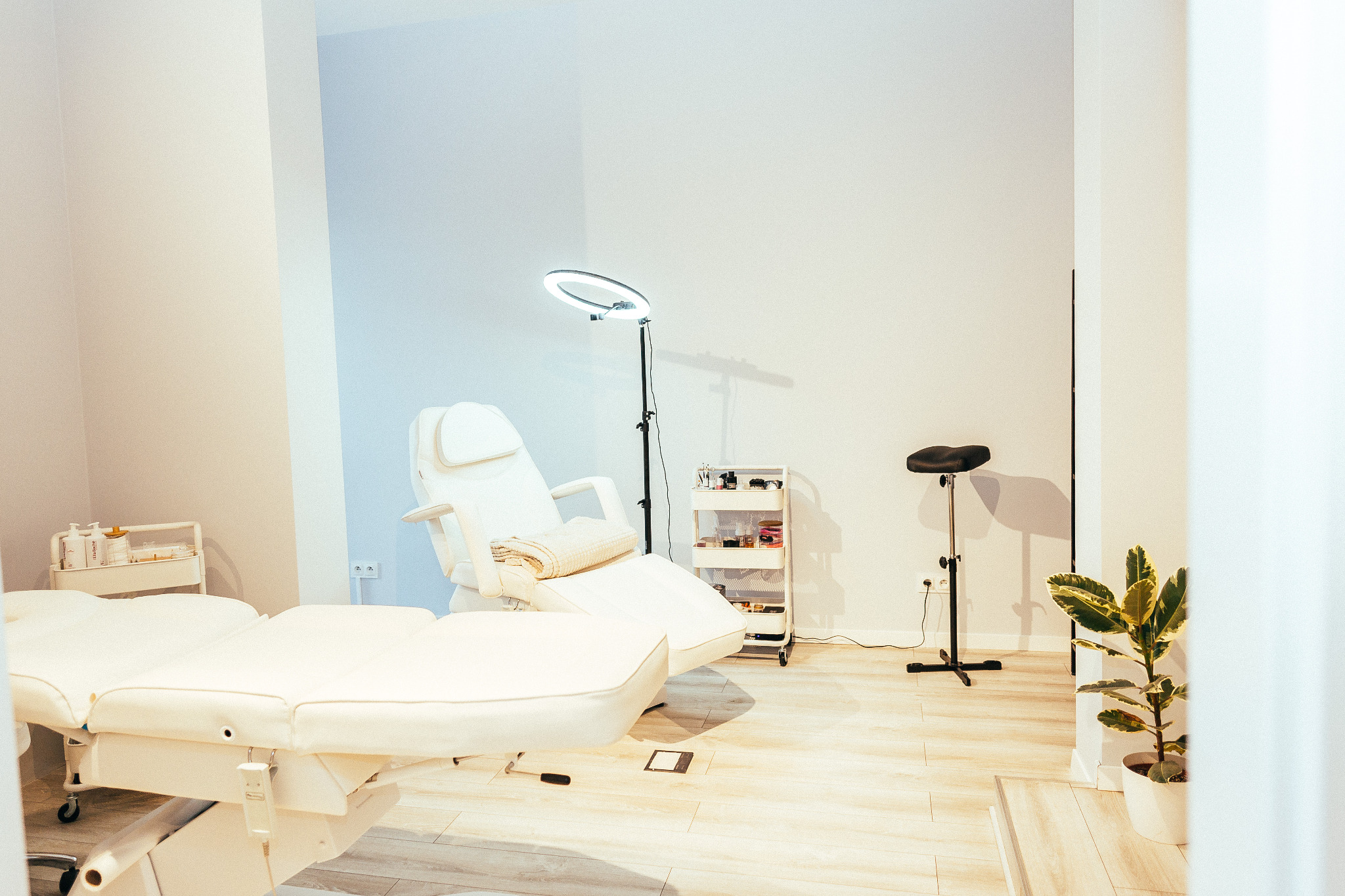 Jasne wnętrze gabinetu z białym fotelem kosmetycznym, lampą pierścieniową, wózkiem z kosmetykami i czarnym stołkiem obok rośliny doniczkowej.