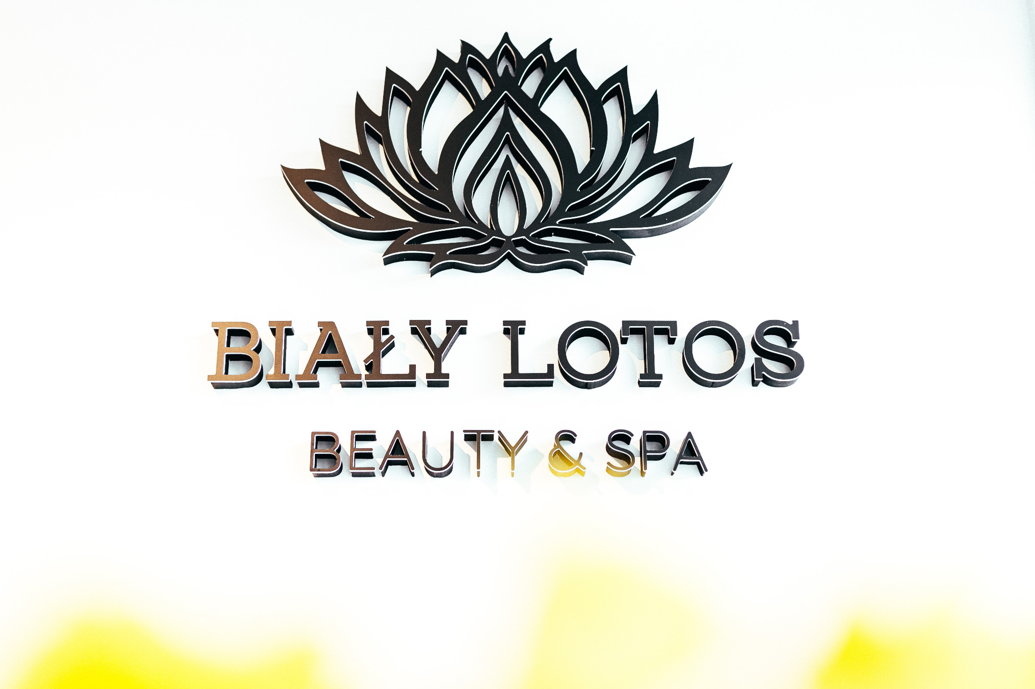 Logo salonu 'Biały Lotos Beauty & Spa' z dekoracyjnym motywem lotosu nad napisem, wykonane z trójwymiarowych liter i elementów, umieszczone na białej ścianie z delikatnym żółtym gradientem u dołu.