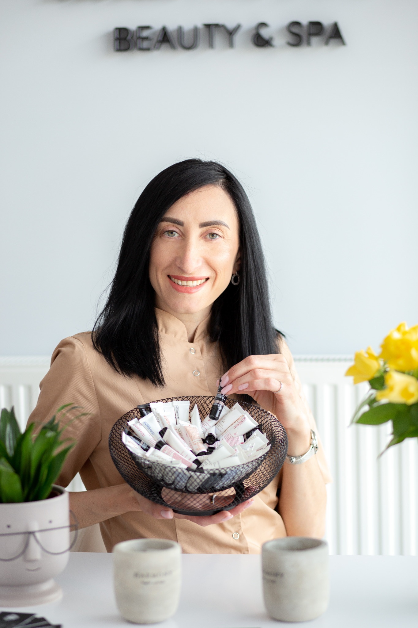 Kobieta w beżowej koszuli trzyma koszyk pełen miniatur kosmetyków, uśmiechając się do kamery, na tle białej ściany z napisem 'BEAUTY & SPA', obok wazon z żółtymi kwiatami i doniczka z zieloną rośliną.