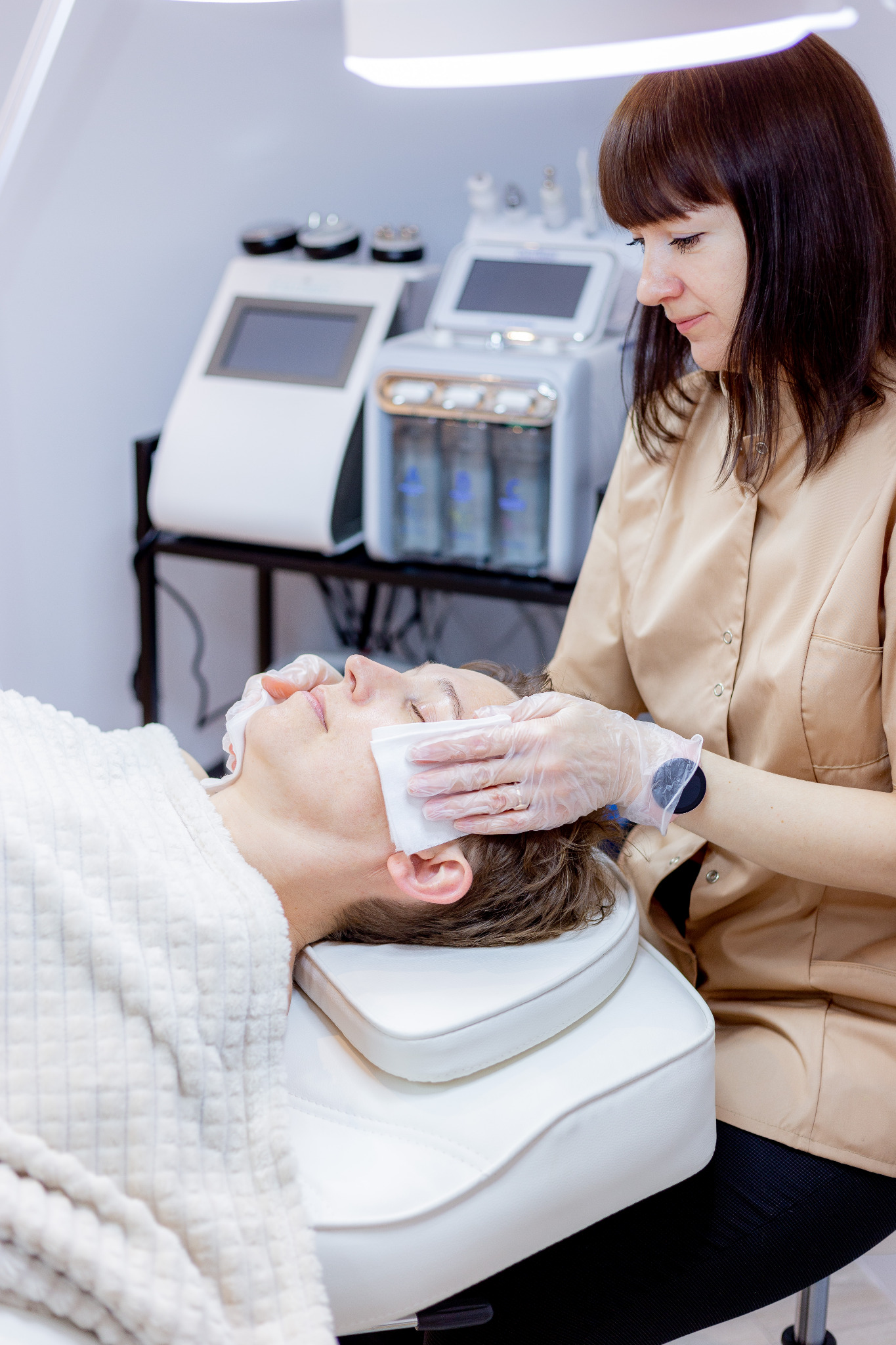 Klientka leży na białym fotelu, a kosmetyczka w beżowej bluzce, w rękawiczkach, przykłada waciki do twarzy. W tle widoczne są urządzenia do zabiegów.