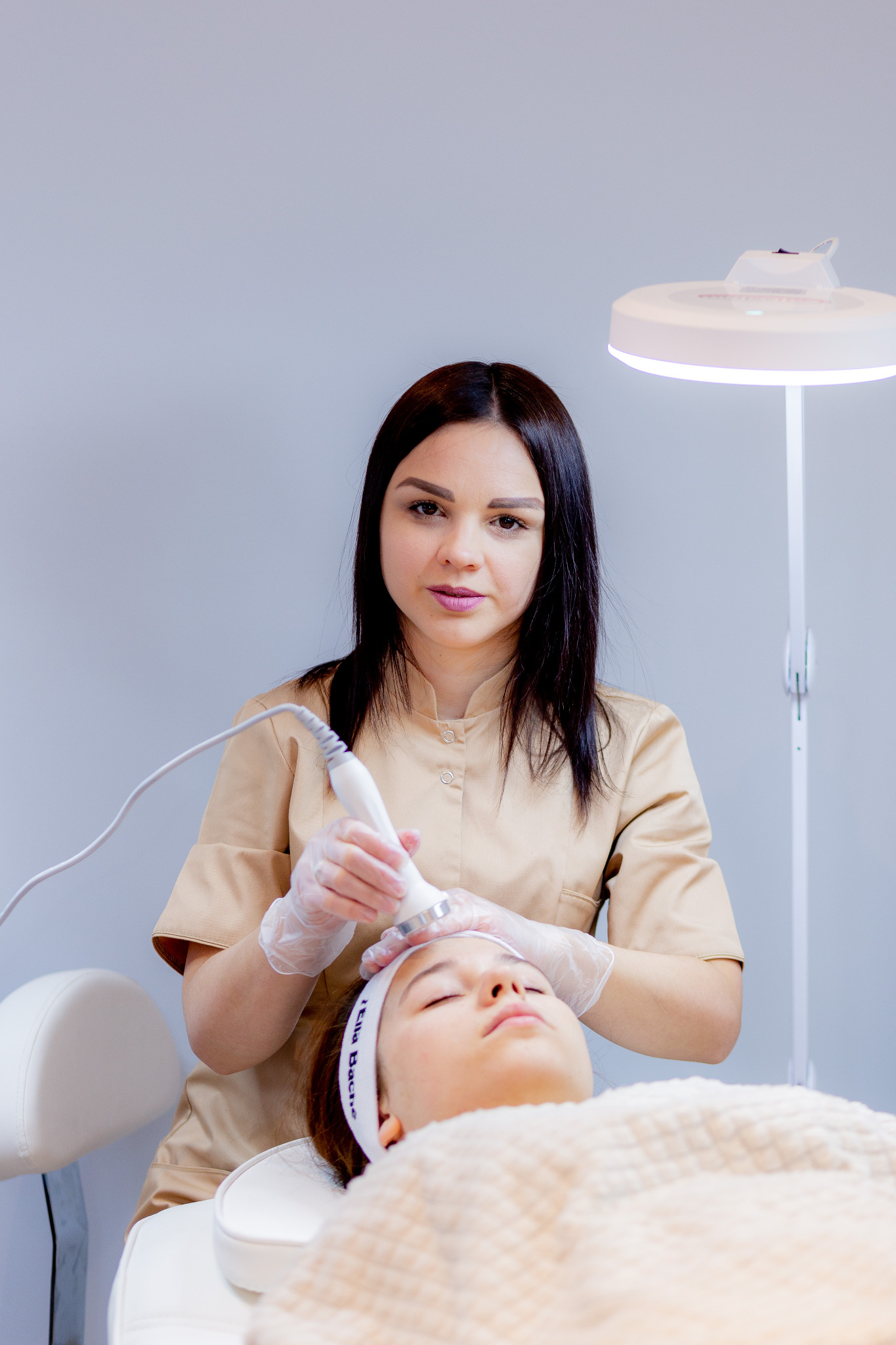 Kobieta w beżowym uniformie wykonuje zabieg na twarzy klientki leżącej na białym fotelu kosmetycznym, używając urządzenia z metalową końcówką, pod jasnym światłem lampy.