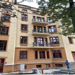 Commodity Renovations - Odnowiona fasada kamienicy z czerwonymi oknami i balkonami, widok z dołu, z elementami rusztowania i pracownikami w tle. Jasna, słoneczna kolorystyka.