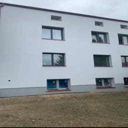 Commodity Renovations - Odnowiona elewacja budynku z nowymi oknami, świeżo po tynkowaniu. Widoczne czerwone rynny i szara podmurówka. Część okien zabezpieczona folią.