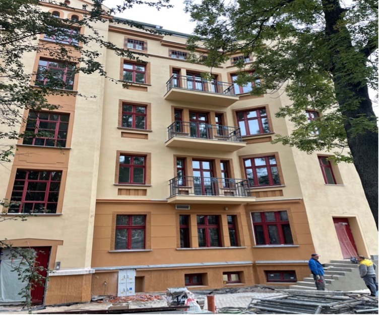 Odnowiona fasada kamienicy z czerwonymi oknami i balkonami, widok z dołu, z elementami rusztowania i pracownikami w tle. Jasna, słoneczna kolorystyka.