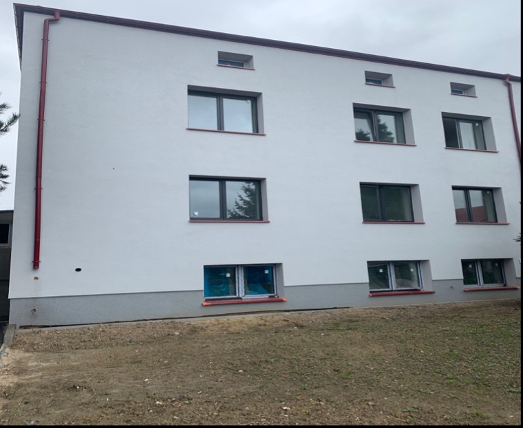 Odnowiona elewacja budynku z nowymi oknami, świeżo po tynkowaniu. Widoczne czerwone rynny i szara podmurówka. Część okien zabezpieczona folią.