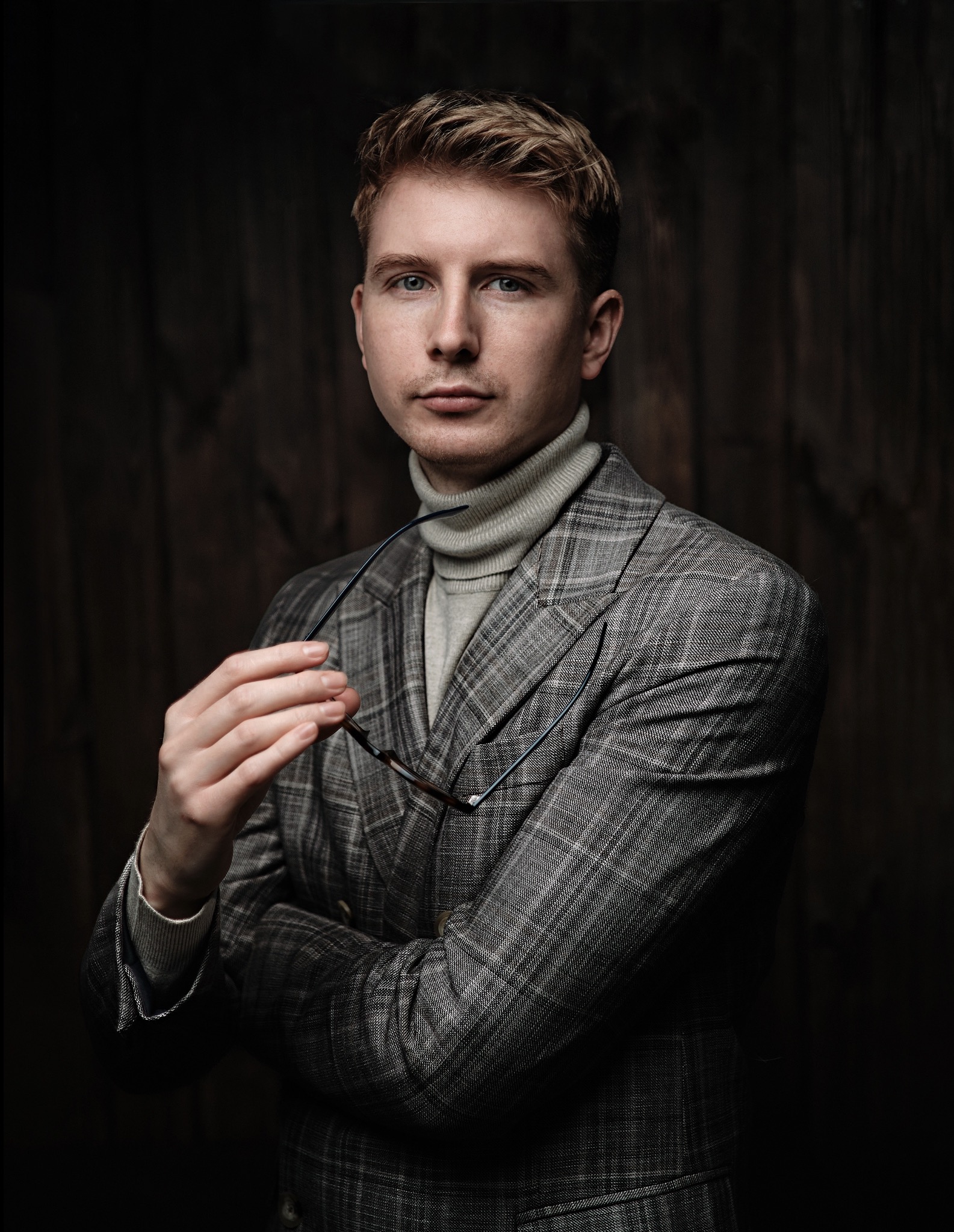 Portret młodego mężczyzny w szarym garniturze w kratę i golfie, trzymającego okulary na tle ciemnego, drewnianego tła.