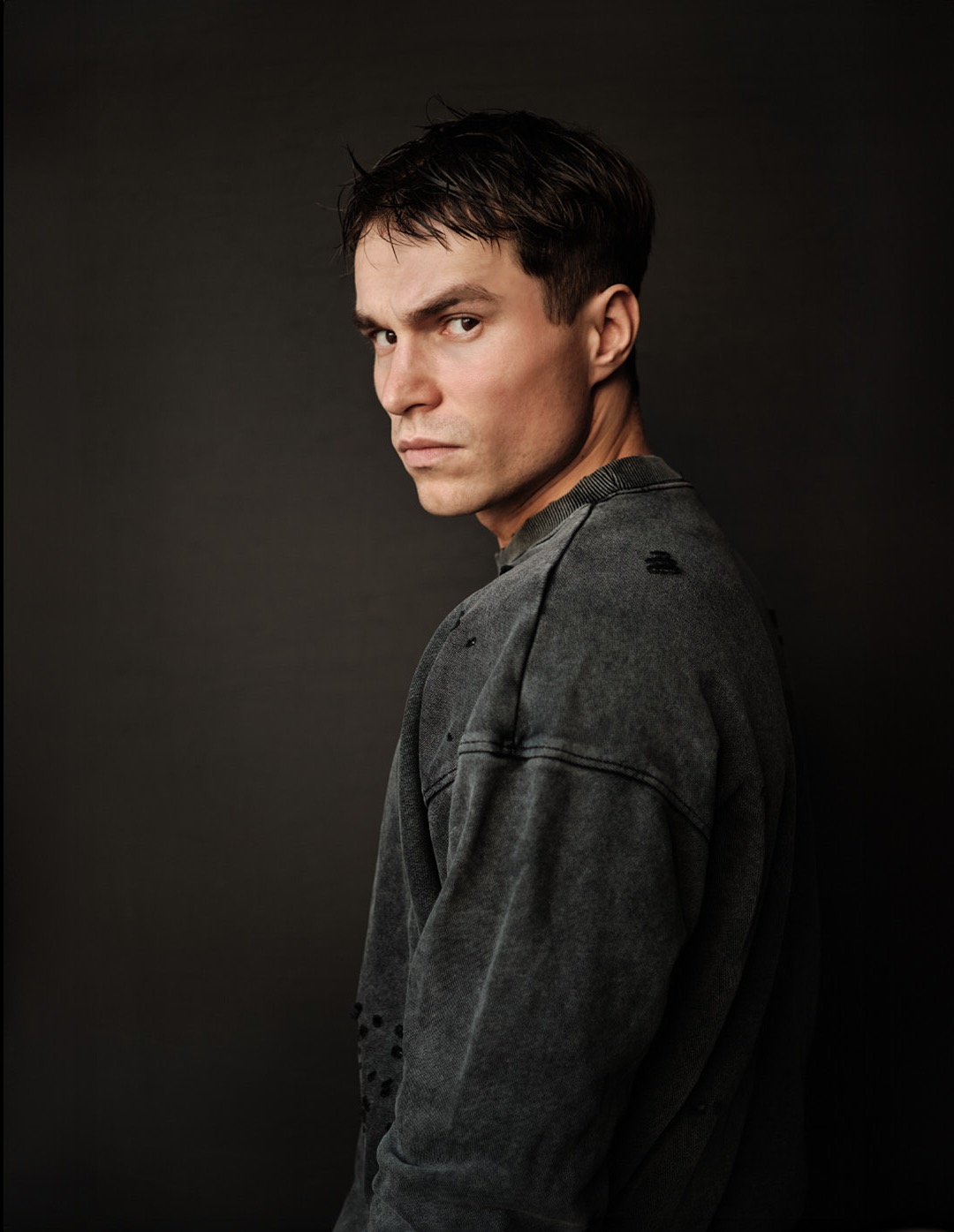 Portret mężczyzny z krótkimi, mokrymi włosami, odwróconego przez ramię, ubrany w szarą bluzę z dziurami na ciemnym tle.