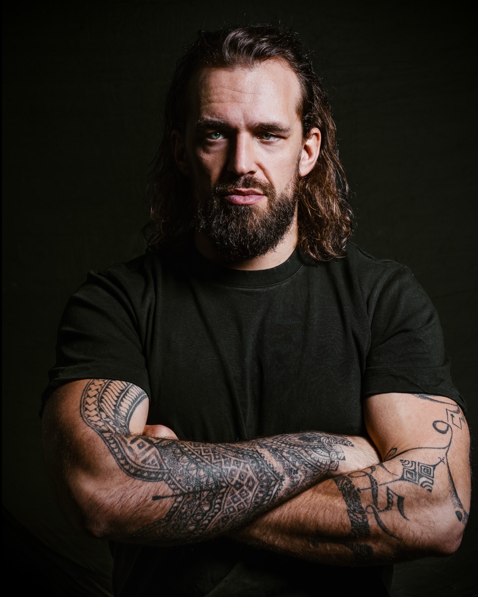 Portret mężczyzny z brodą i długimi włosami, z tatuażami na ramionach, pozującego na ciemnym tle.
