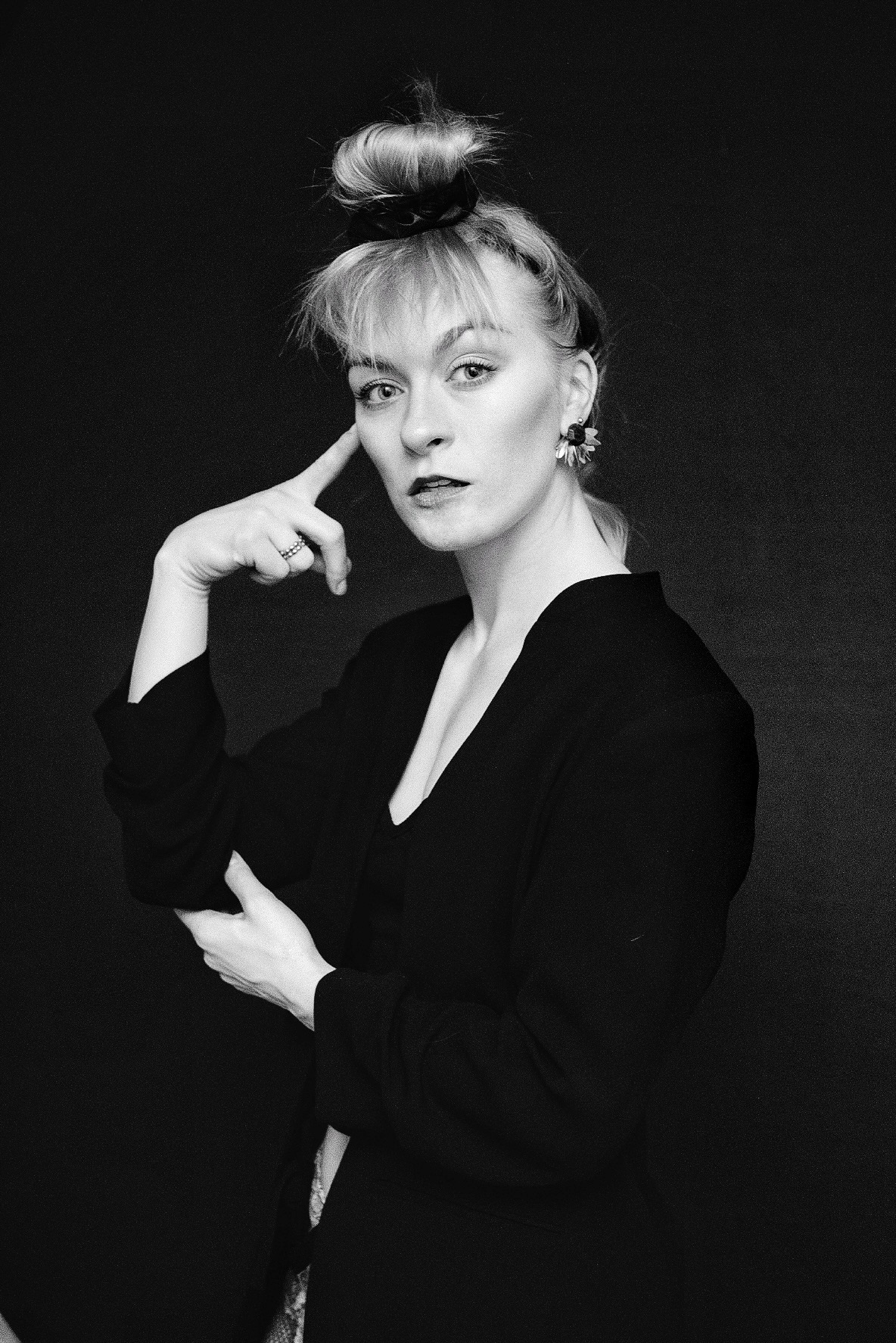 Czarno-białe portretowe ujęcie kobiety z blond włosami upiętymi w kok, z ręką przy skroni, w ciemnym ubraniu i z kwiatowymi kolczykami na tle ciemnego tła.